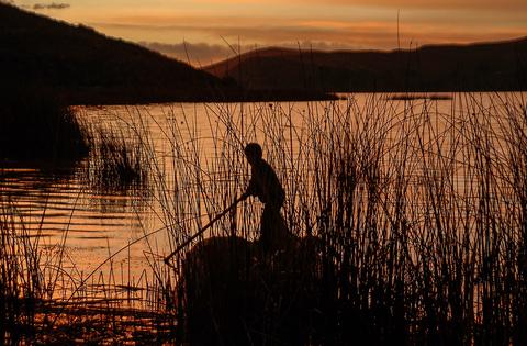 Un pescador navega en un lago al atardecer, rodeado de cañas y reflejos dorados en el agua.