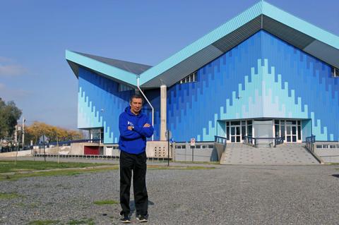 Un hombre de pie frente a un moderno edificio con un diseño triangular y colores azul y gris.
