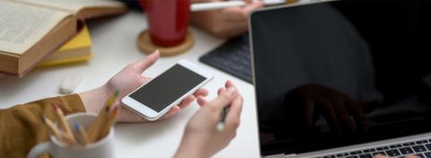 Una mano sostiene un teléfono móvil sobre una mesa con una laptop y una taza de café al fondo.