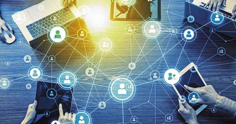 La imagen muestra una mesa con diversas personas usando dispositivos electrónicos interconectados.