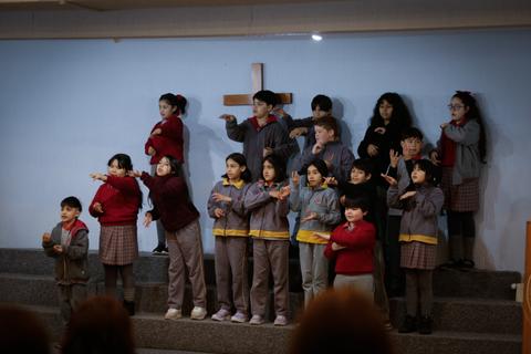 Un grupo de niños en un acto artístico en un escenario con una cruz al fondo.
