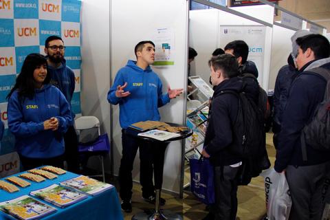 Un grupo de personas interactúa en un stand universitario durante un evento.