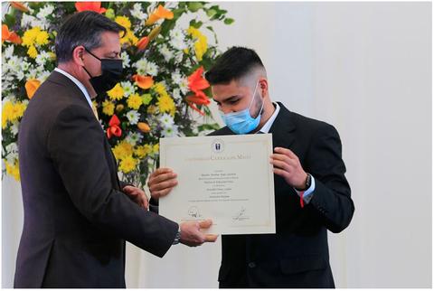 Un joven recibe un certificado de manos de un hombre mayor en un evento formal con flores de fondo.
