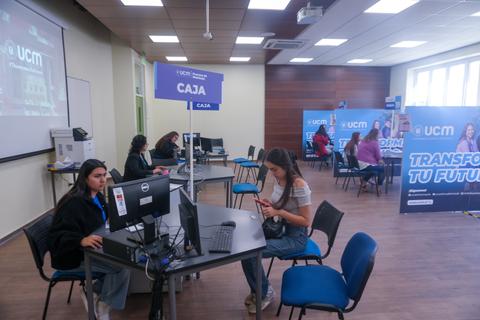 La imagen muestra un aula con varias personas sentadas frente a computadoras y un cartel que dice 'CAJA'.