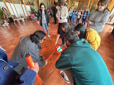Un grupo de personas participa en una actividad grupal utilizando pelotas y tubos de colores en un espacio cerrado.