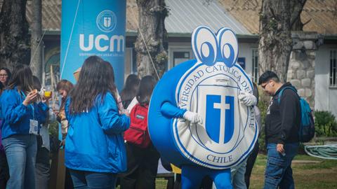 Un grupo de personas se reúne alrededor de una mascota de la Universidad Católica de Chile en un evento al aire libre.