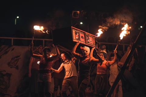 Un grupo de personas sostiene un ataúd decorado mientras sostienen antorchas en una representación teatral nocturna.