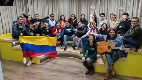Un grupo de jóvenes sonrientes pose para la foto sosteniendo una bandera de Colombia en un ambiente alegre y comunitario.