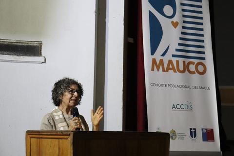 Una mujer está hablando en un evento, frente a un cartel de MAUCO que representa una cohorte poblacional.