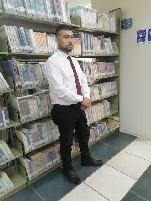 Un hombre vestido formalmente de pie junto a estanterías llenas de libros en una biblioteca.