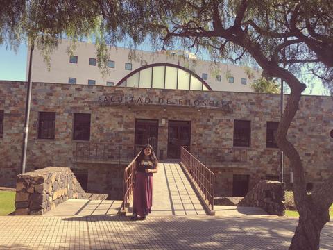 Una estudiante posa frente a la Facultad de Filosofía, en un entorno natural con árboles.