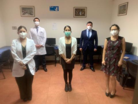 Un grupo de cinco personas, vestidas de manera formal y con mascarillas, se encuentran de pie en una sala con mobiliario sencillo.