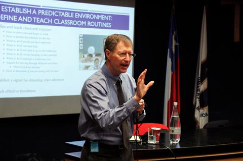 Un hombre presenta una charla ante un público, mientras señala una pantalla con información educativa.