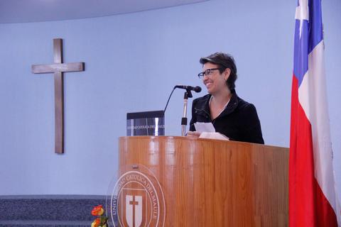 Una mujer sonriente habla desde un podio en un evento en una institución con una cruz en la pared y la bandera de Chile al fondo.