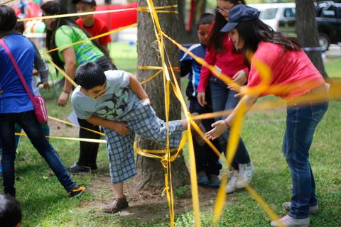 Un grupo de niños juega alrededor de un árbol, atravesando cintas de colores en un parque.