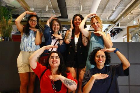 Un grupo de seis mujeres posando con las manos arriba en una oficina moderna.