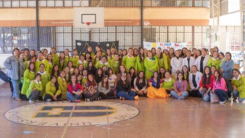 Un grupo grande de mujeres reunidas en un gimnasio, todas con vestimenta colorida y sonrisas en sus rostros.