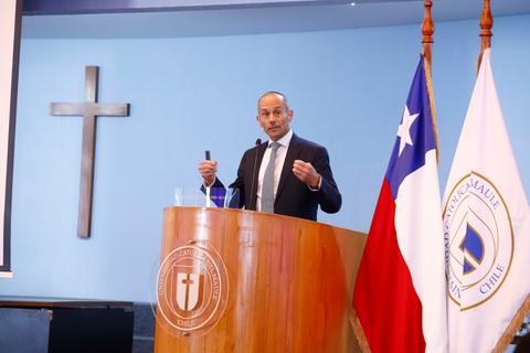 Un hombre dando una charla en un auditorio con una cruz y banderas chilenas de fondo.