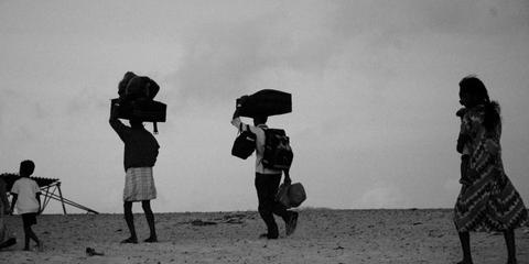 Un grupo de personas camina por la playa llevando maletas y objetos sobre sus cabezas.
