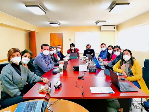 Un grupo de personas con mascarillas se encuentra reunido en una sala de trabajo con computadoras y un proyector.