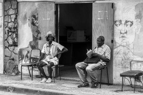 Dos personas sentadas frente a un local, una de ellas leyendo un libro y la otra mirando hacia el horizonte.