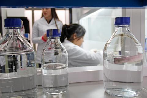 En la imagen se ven frascos de laboratorio con líquidos y una persona trabajando al fondo.