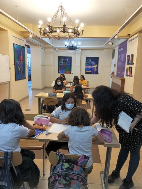 Un grupo de estudiantes trabaja en un salón de clases con una profesora ayudando a una de ellas.