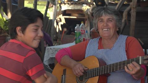 Un niño sonriente escucha a una mujer mayor que toca la guitarra en un ambiente rural.