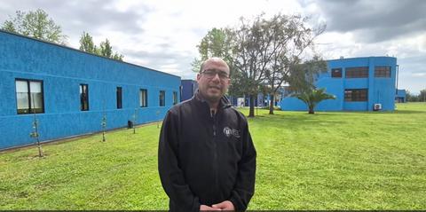 Un hombre con chaqueta negra posa frente a un edificio azul rodeado de césped verde.