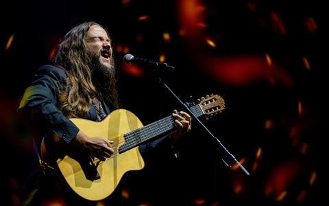 Un músico con cabello largo canta apasionadamente mientras toca su guitarra en un escenario iluminado.