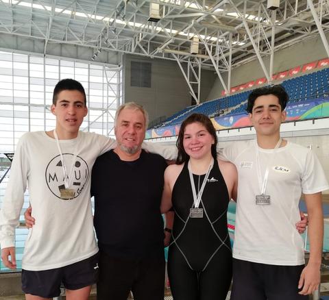 Un grupo de cuatro personas, incluido un entrenador, posan felices con medallas tras una competencia de natación.
