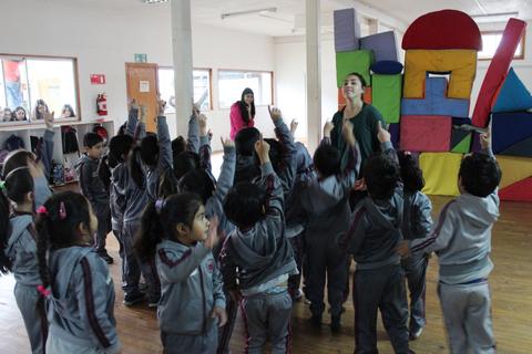 Un grupo de niños en un salón levantando la mano mientras una mujer les habla.