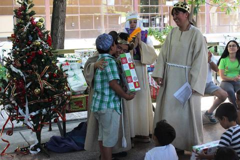 Una celebración navideña donde un niño recibe un regalo de manos de personas disfrazadas y hay un árbol de Navidad decorado.
