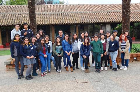 Un grupo grande de jóvenes posando en un espacio abierto con vegetación y un edificio al fondo.