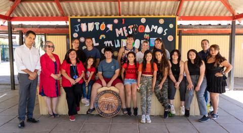 Un grupo de estudiantes y adultos posan juntos frente a un mural escolar que dice 'Juntos mejoramos mi mundo'.