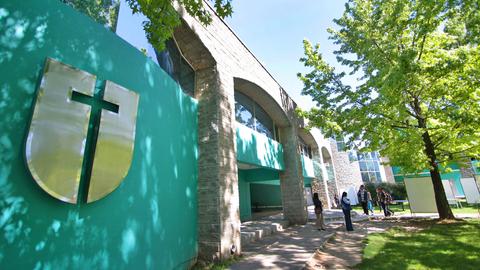La imagen muestra un edificio moderno con un gran logo en la pared y personas caminando por el exterior.