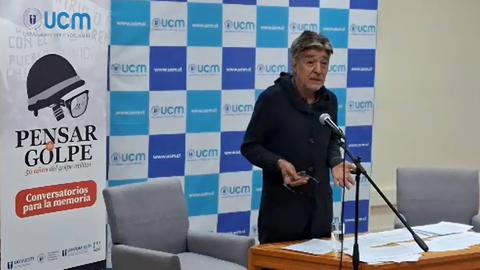Un hombre está hablando en un evento sobre la memoria histórica en un salón con decoraciones institucionales.