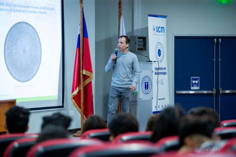 Un hombre está dando una presentación en un auditorio con banderas y una pantalla de proyección detrás.
