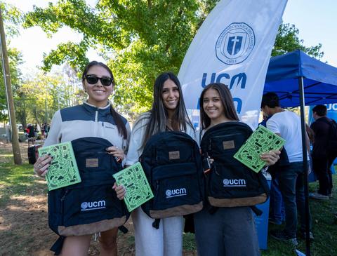 Tres jóvenes posan felices con mochilas y cuadernos en un evento al aire libre.