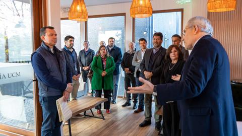 Un grupo de personas asiste a una reunión en un espacio cerrado, mientras un hombre habla de pie frente a otro.