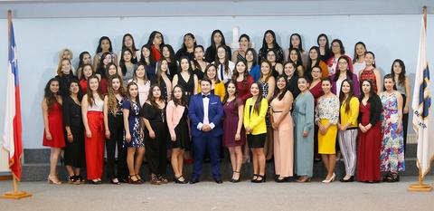 Un grupo grande de mujeres de pie en un evento formal, posando junto a un hombre vestido de traje en un escenario.
