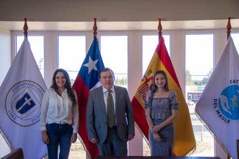 Tres personas posan juntas en un entorno institucional con banderas de Chile y España.