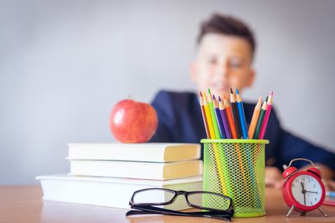 Un niño sonriente está sentado frente a una mesa con libros, un manzana, lápices de colores y un reloj despertador.