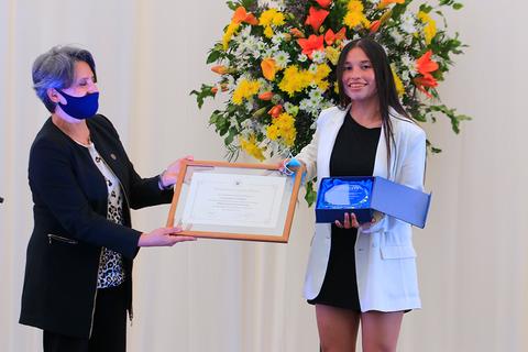Una joven sonriente recibe un premio de una mujer en un evento formal, frente a un fondo decorado con flores.