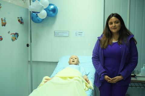 Una mujer vestida de morado está de pie junto a un maniquí en una cama de hospital.