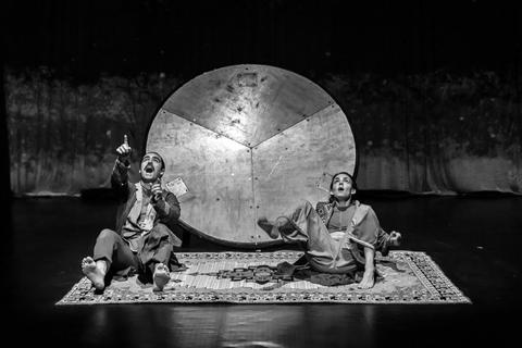 Dos actores en una actuación teatral, sentados sobre una alfombra, uno señala hacia arriba mientras el otro se encuentra en una posición relajada.