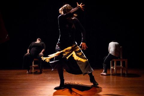 Una mujer baila con una falda que gira en un fondo oscuro mientras otras dos personas están sentadas y no miran hacia el frente.