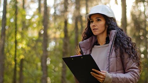 Una mujer con casco observa atentamente un paisaje forestal mientras sostiene una hoja de papel.