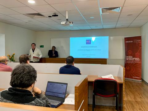 Una presentación en un aula con varias personas asistiendo y un proyector mostrando una diapositiva.