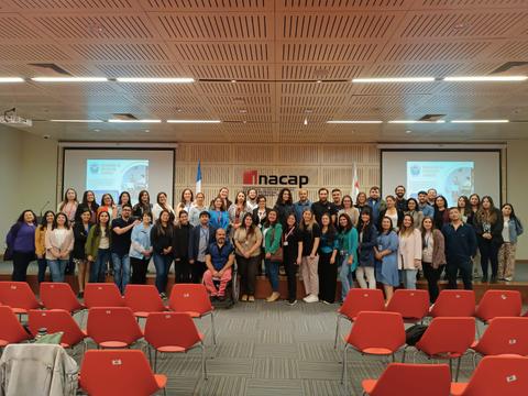 Un grupo grande de personas posa para una foto en un evento en un salón con sillas rojas y pantallas al fondo.